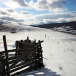 A rustic split rail fence with wood members stacked end over end stretches away from view, over rolling snow covered hills towards mountains beyond on a sunny cloud filled day.