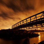 Bright, cloudy skies, Summer nights and site lighting located at steel pedestrian bridge on the Norfolk Naval base create an image with a silhouette for the bridge and rocks with bright orange for the clouds and water beneath the bridge.