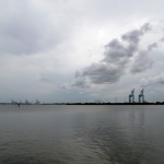 Gray, still water with reflections of industrial shipping cranes fills the lower third of a photograph while the top two-thirds are filled with white and gray clouds with sunlight streaming from behind two large clouds.