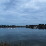 A view of the LaFayette River Bridge during the blue hour as seen from within the Larchmont neighborhood in Norfolk, Virginia. Clouds completely cover the sky, the warm yellow from the street lights on the bridge are reflected in the medium blue hue of the water.