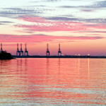 Industrial cranes sit on the horizon at the edge between water and sky. The sun is setting so the sky is glowing bright pink and the color is reflected in the water with the edges of the image appearing a silvery blue.
