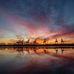 Landscape of Industrial shipping cranes reflected at water’s edge during vivid sunset with streaks of clouds across the sky.