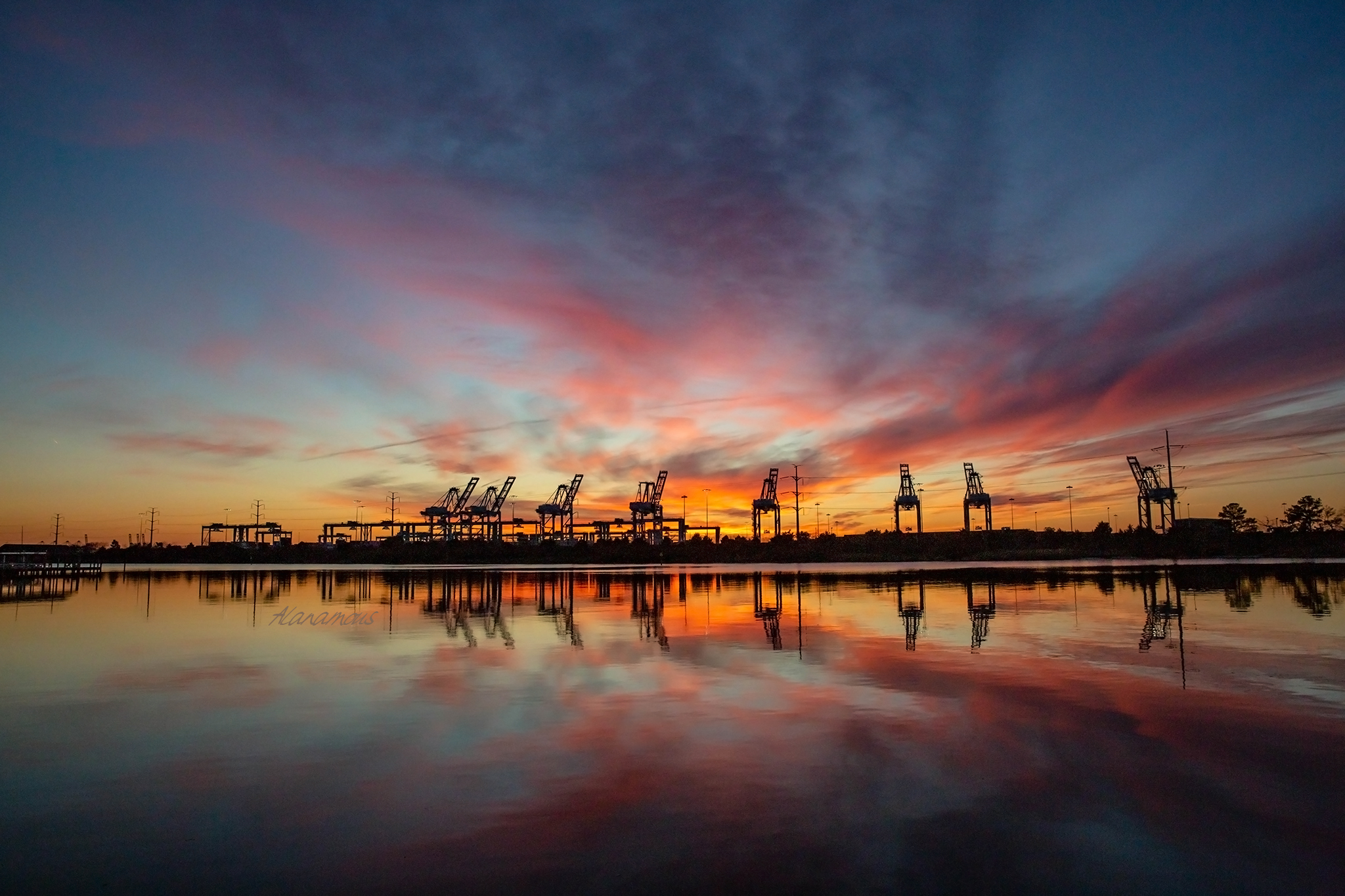 Landscape of Industrial shipping cranes reflected at water’s edge during vivid sunset with streaks of clouds across the sky.